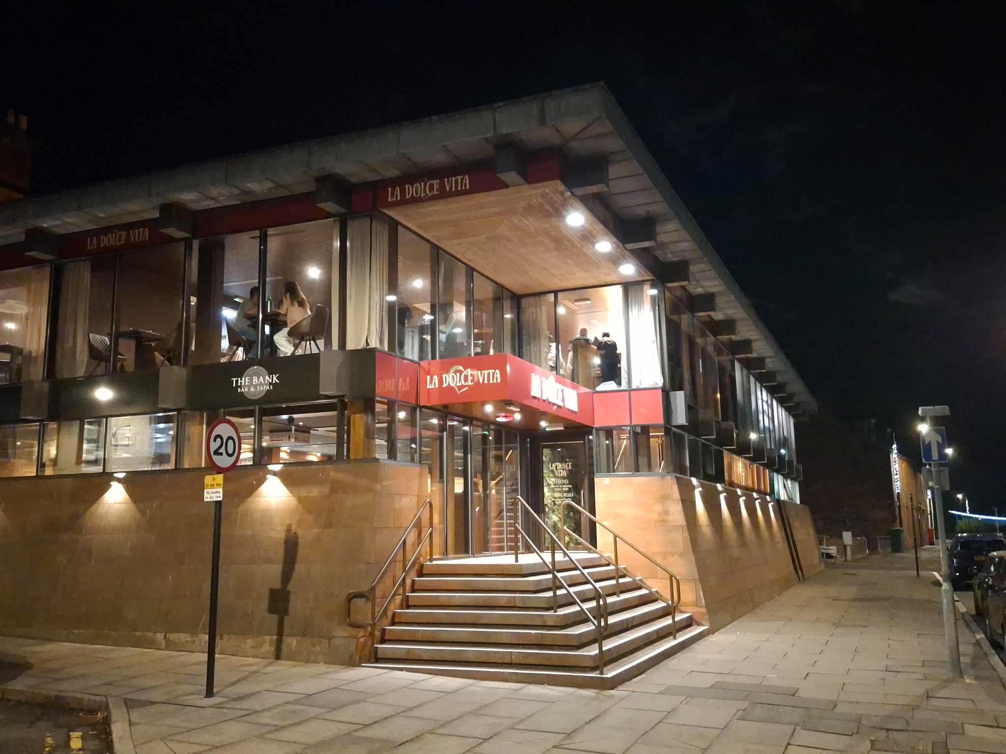 The building at night from the corner, with The Bank Bar & Tapas signed on the lower floor and La Dolce Vita above it